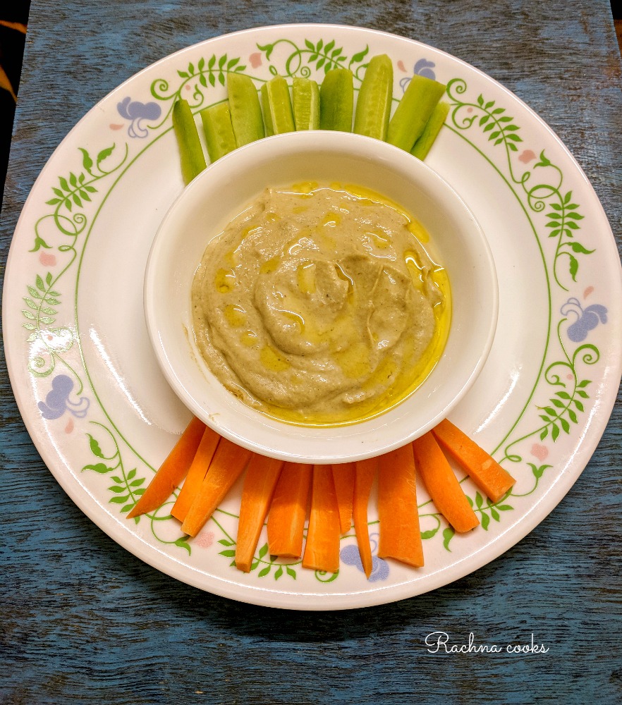 Baba ganoush dip in a white bowl with slices of carrot and cucumber