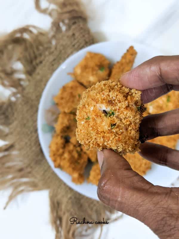 Close up of one golden brown chicken nugget held in hand. Blurred background shows a white plate with more nuggets.