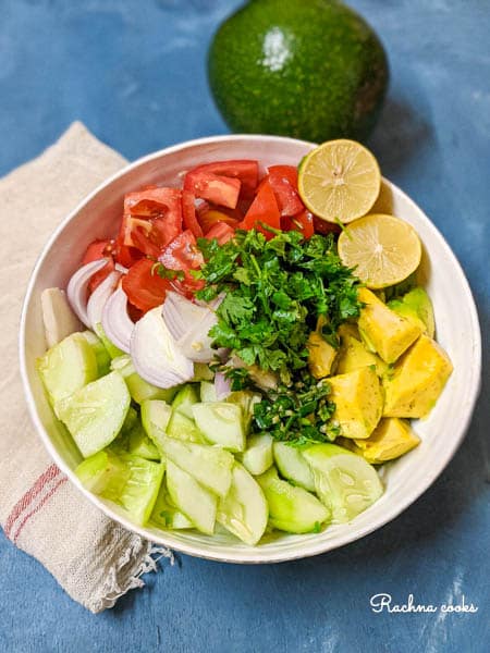 In a white salad bowl are chopped cucumber, avocado, tomato, sliced onion, cilantro and jalapeno. There is a blue background with a white napkin and an avocado.