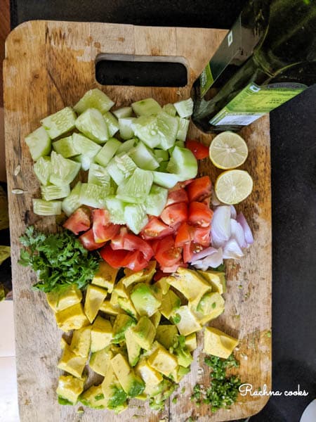 On a chopping board there are chopped cucumber, tomato, avocado, cilantro, onion, a lime and a bottle of olive oil.