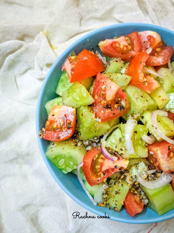 A top shot of half a blue bowl showing juicy pieces of red tomatoes, green cucumber and onion slices with a glistening dressing on a white background.