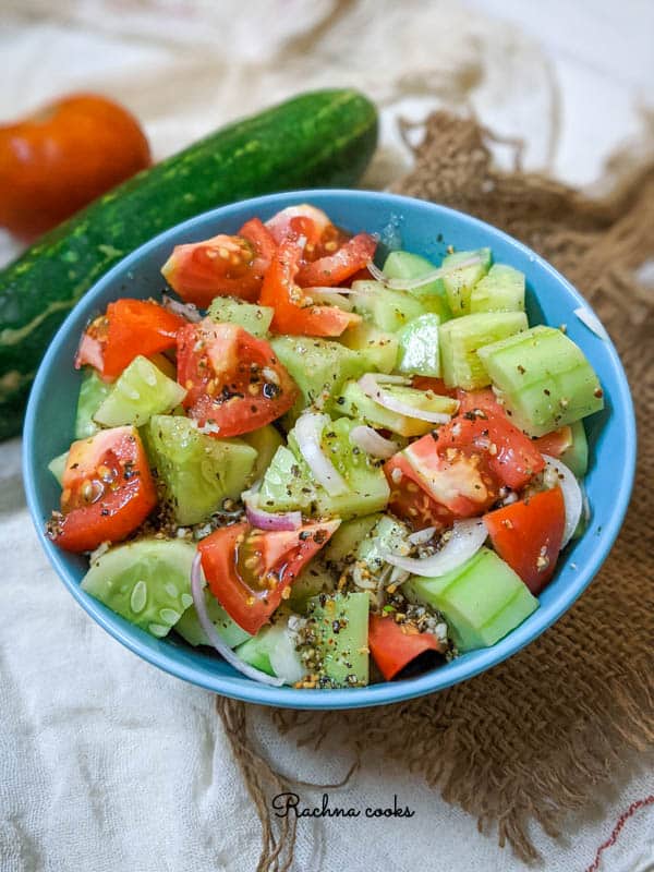 A blue bowl showing glistening pieces of cucumber, tomato and sliced onions with dressing. In the background you can see tomato and cucumber