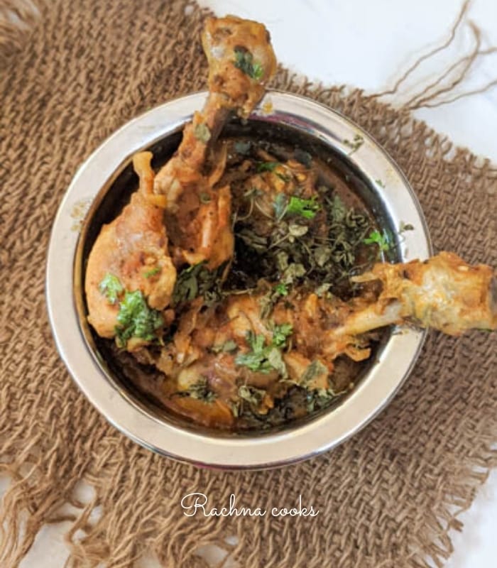Top shot of a  round pan shows chicken drumsticks coated in rich red curry with a garnish of dry fenugreek and cilantro on brown mat on a white background.