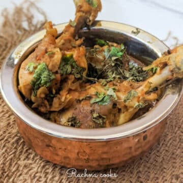 Top shot of a round pan shows chicken drumsticks coated in rich red curry with a garnish of dry fenugreek and cilantro on brown mat on a white background.
