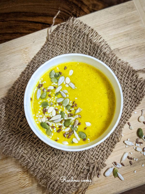 Top shot of yellow pumpkin soup garnished with pumpkin, sesame and melon seeds in a white bowl on a brown mat and light brown background.