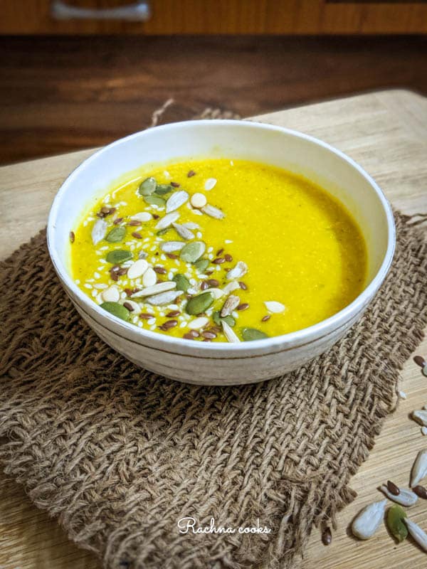 Side shot of yellow pumpkin soup garnished with pumpkin, sesame and melon seeds in a white bowl on a brown mat and light brown background.