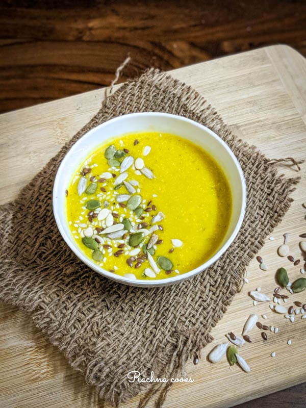 Side shot of yellow pumpkin soup garnished with pumpkin, sesame and melon seeds in a white bowl on a brown mat and light brown background.