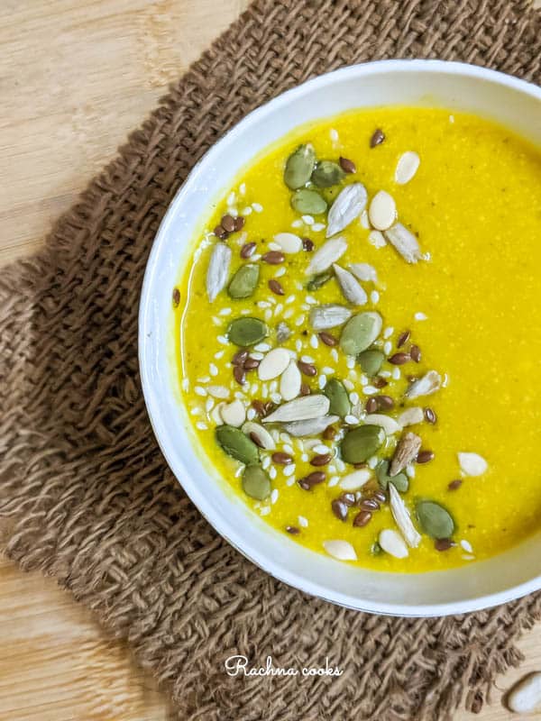 Half Top shot of yellow pumpkin soup garnished with pumpkin, sesame and melon seeds in a white bowl on a brown mat and light brown background.