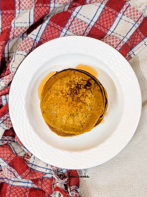 Top shot of Bright yellow orange pumpkin pancakes that look golden brown with honey on top and stacked together on a white plate with a red and white napkin in background.