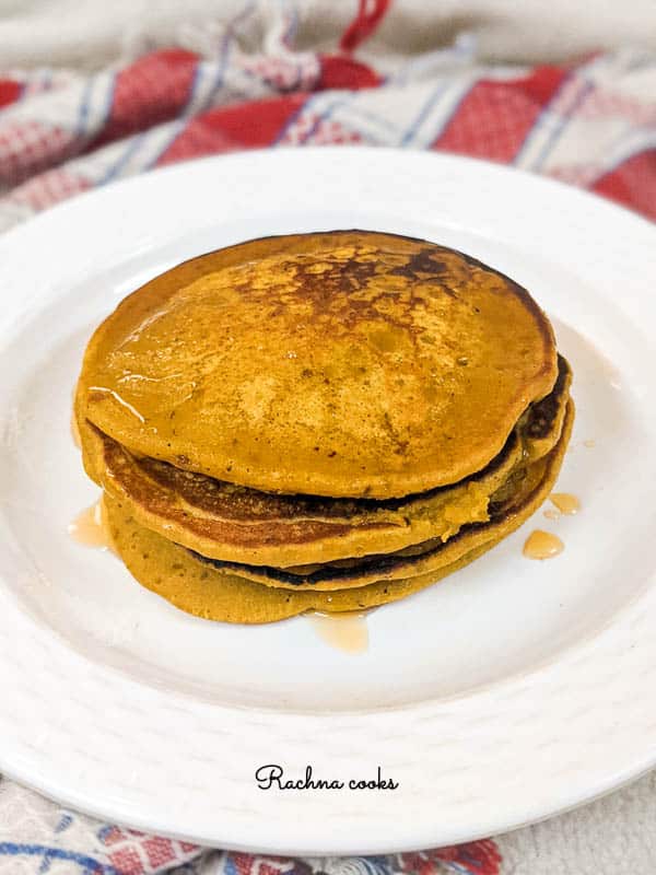 Bright yellow orange pumpkin pancakes that look golden brown with honey on top and stacked together on a white plate with a red and white napkin in background.