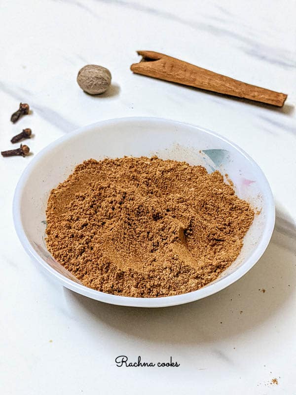 Warm brown pumpkin pie spice in a white bowl with pink flower pattern. Background is white with grey streaks with nutmeg, clove and cinnamon spices.