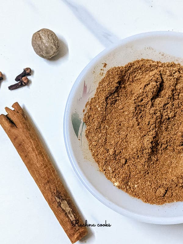 Warm brown pumpkin pie spice in a white bowl with pink flower pattern. Background is white with grey streaks with nutmeg, clove and cinnamon spices.