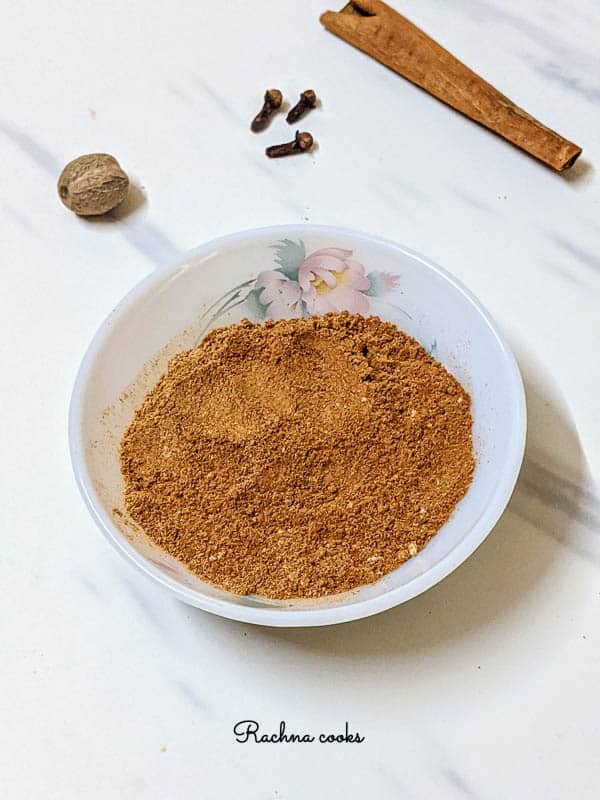 Warm brown pumpkin pie spice in a white bowl with pink flower pattern. Background is white with grey streaks with nutmeg, clove and cinnamon spices.