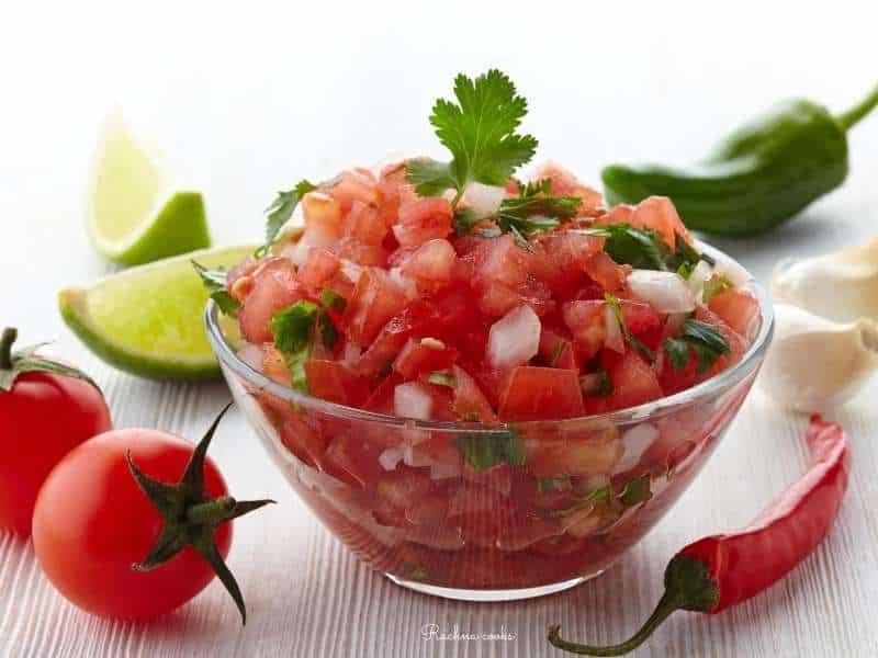 Fresh tomato salsa in a transparent bowl with tomatoes, jalapenos and limes in the background