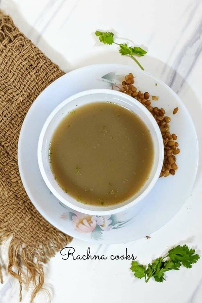 A thin horsegram soup in a round bowl on a white plate with cooked horsegram. This is on a brown mat background.