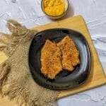 Two golden breaded tilapia fillets after air frying on a black plate with a bowl of dip