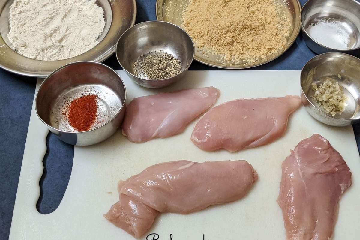 4 raw chicken tenders on a white chopping board with cayenne, minced garlic, pepper, salt, breadcrumbs, flour in shallow bowls and plates.