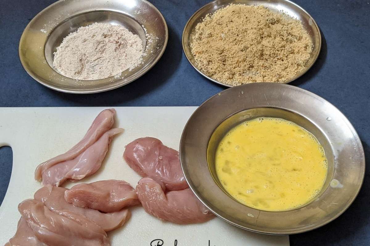 3 shallow bowls having beaten eggs, breadcrumbs with spices and flour along with raw chicken tenders on a white board.