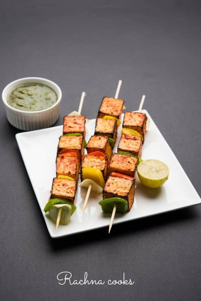 Skewers of paneer tikka with peppers on a white plate served with green chutney and lemon.