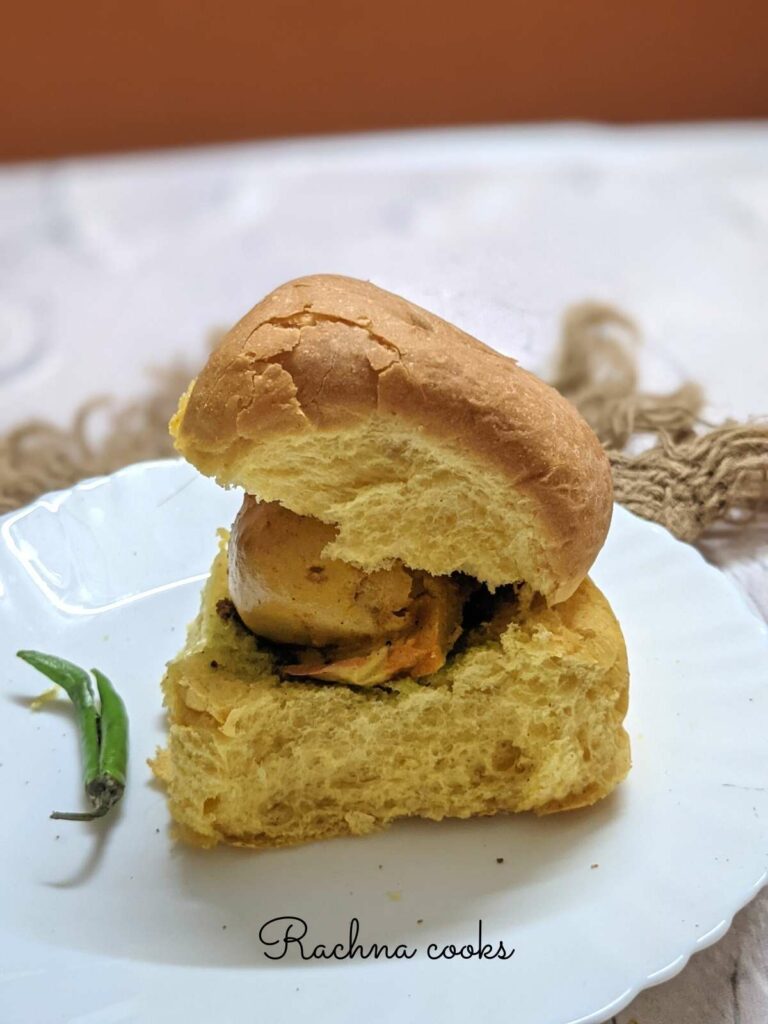 Vada pav on a white plate with a green chilli on the side.