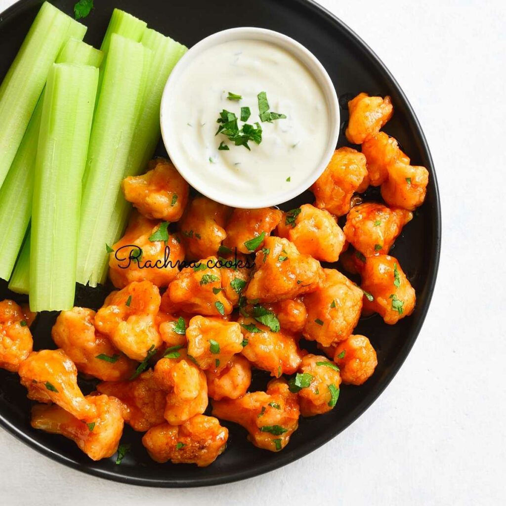 Delicious air fried buffalo cauliflower wings on a black plate with celery on the side and a white dressing in a white bowl.