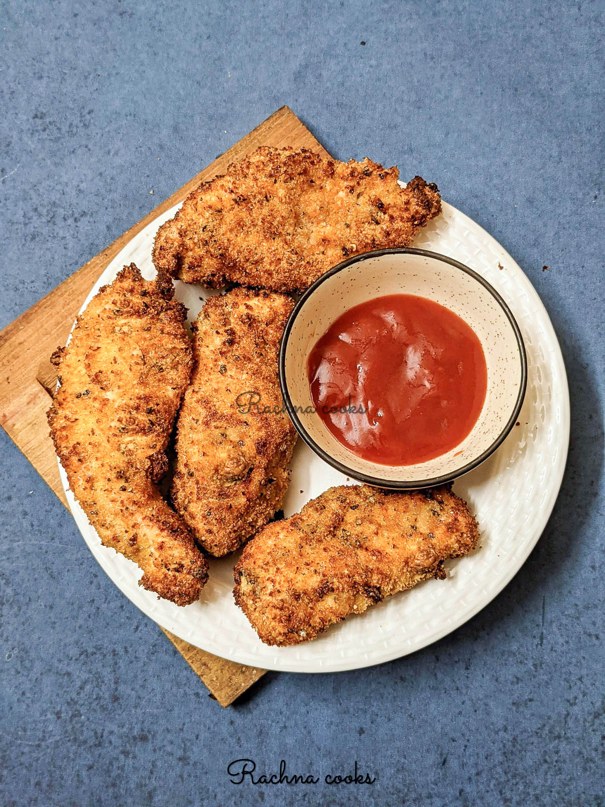4 air fried chicken tenders in a white plate with a bowl of ketchup. This is kept on a slate grey background.