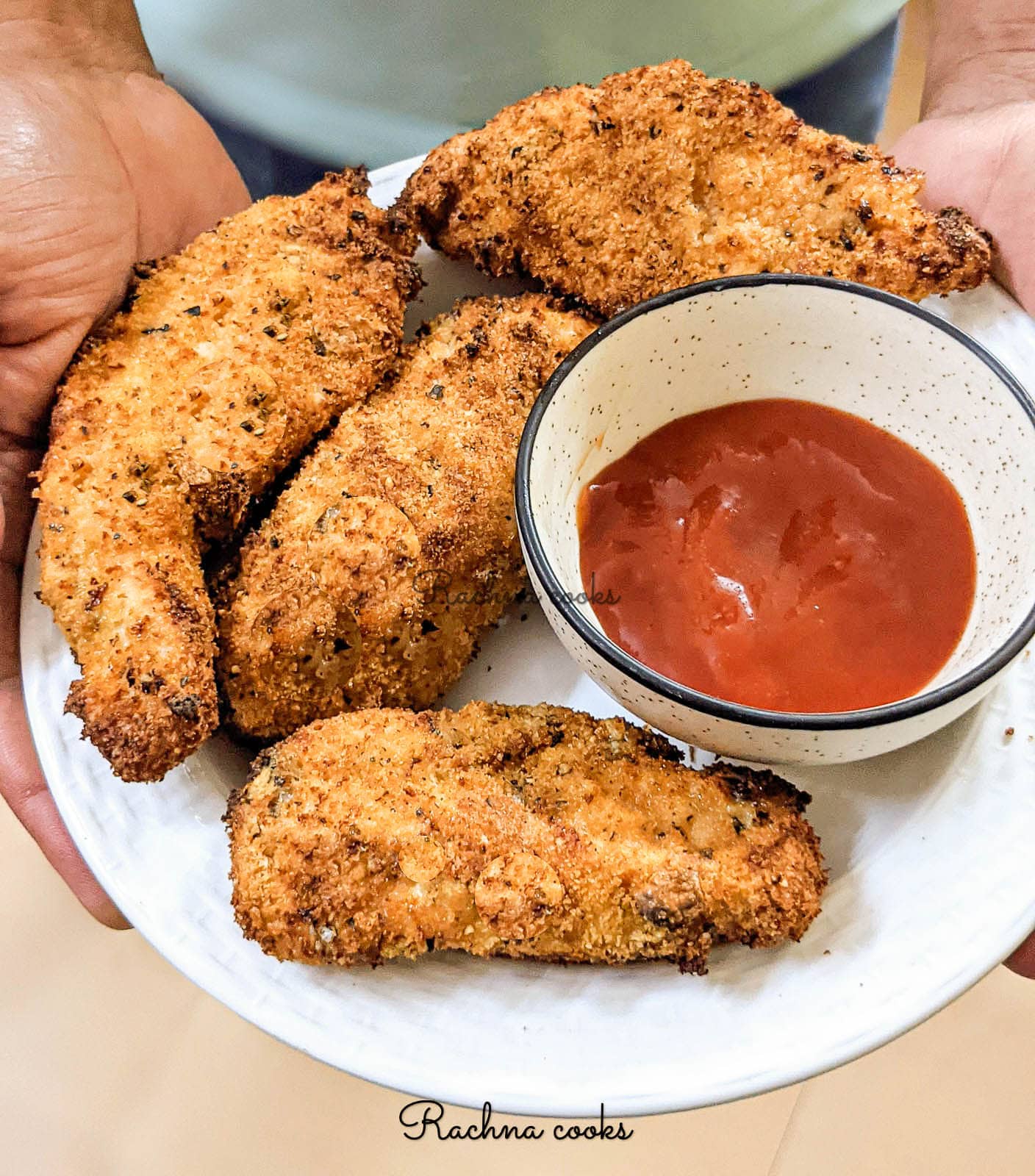 4 air fried chicken tenders in a white plate with a bowl of ketchup. This is kept on a slate grey background.