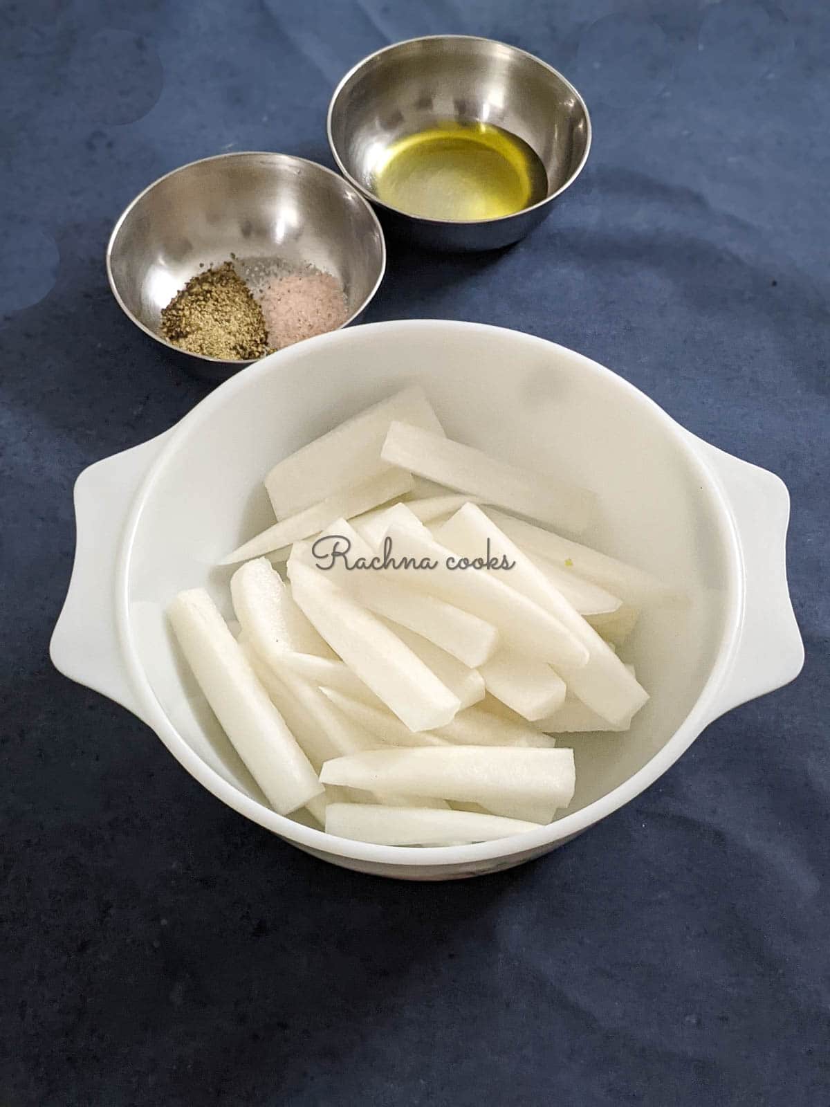 Peeled parsnip fries in a white bowl with pink salt and pepper in a steel bowl and olive oil in another steel bowl.