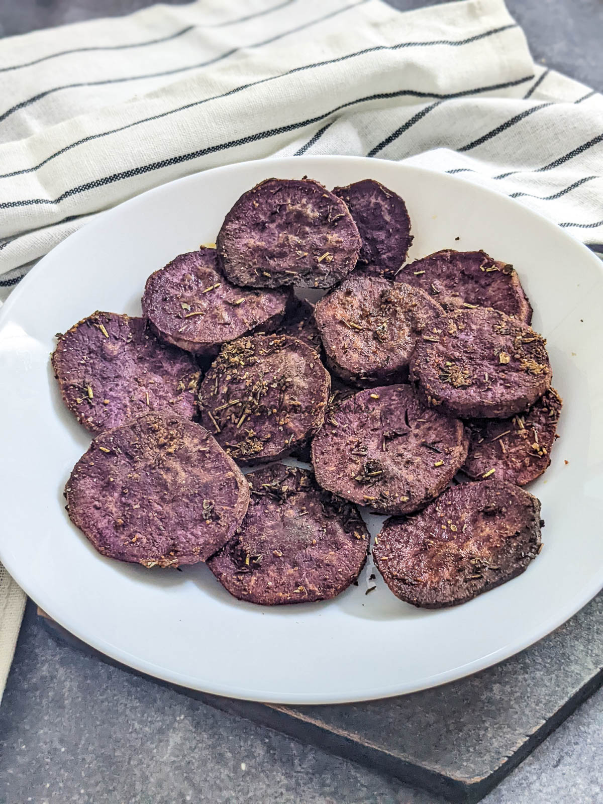 Medium sliced purple sweet potato slices marinated in seasoning and air fried, served on a white plate with a grey background and a napkin visible.
