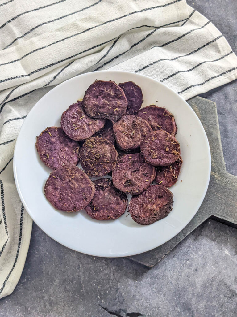 Medium sliced purple sweet potato slices marinated in seasoning and air fried, served on a white plate with a grey background and a napkin visible.