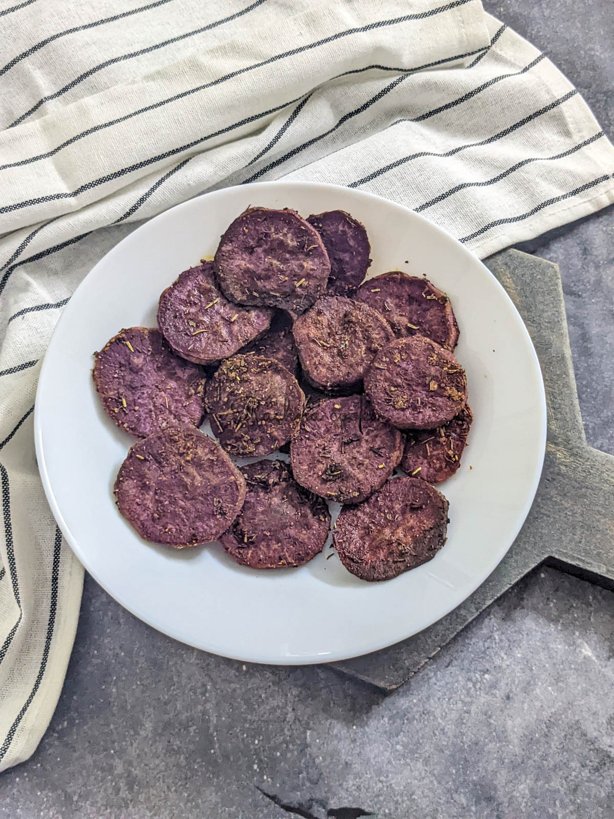 Medium sliced purple sweet potato marinated in seasoning and air fried, served on a white plate with a grey background and a napkin visible.