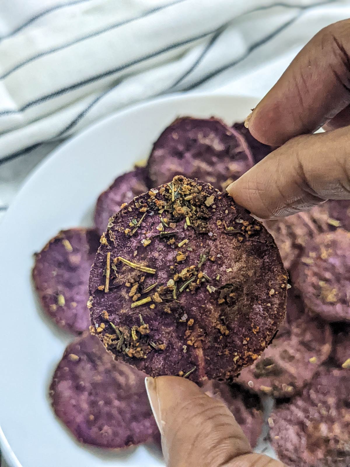 One Medium sliced purple sweet potato slice in close up  with seasoning and air fried, held in fingers with other slices blurred in the background.