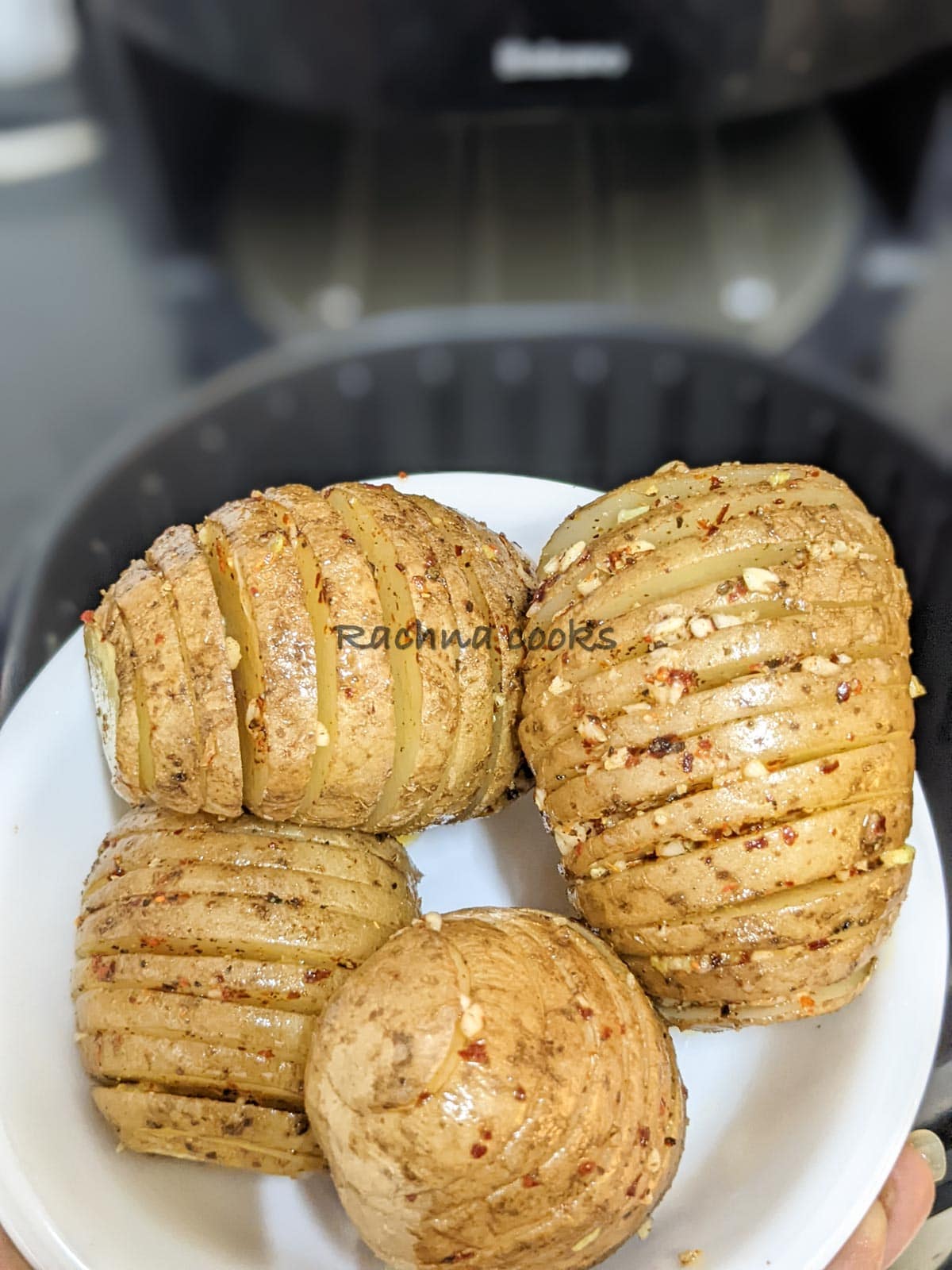 Four hasselback potatoes brushed with seasoning being put into air fryer basket for air frying.
