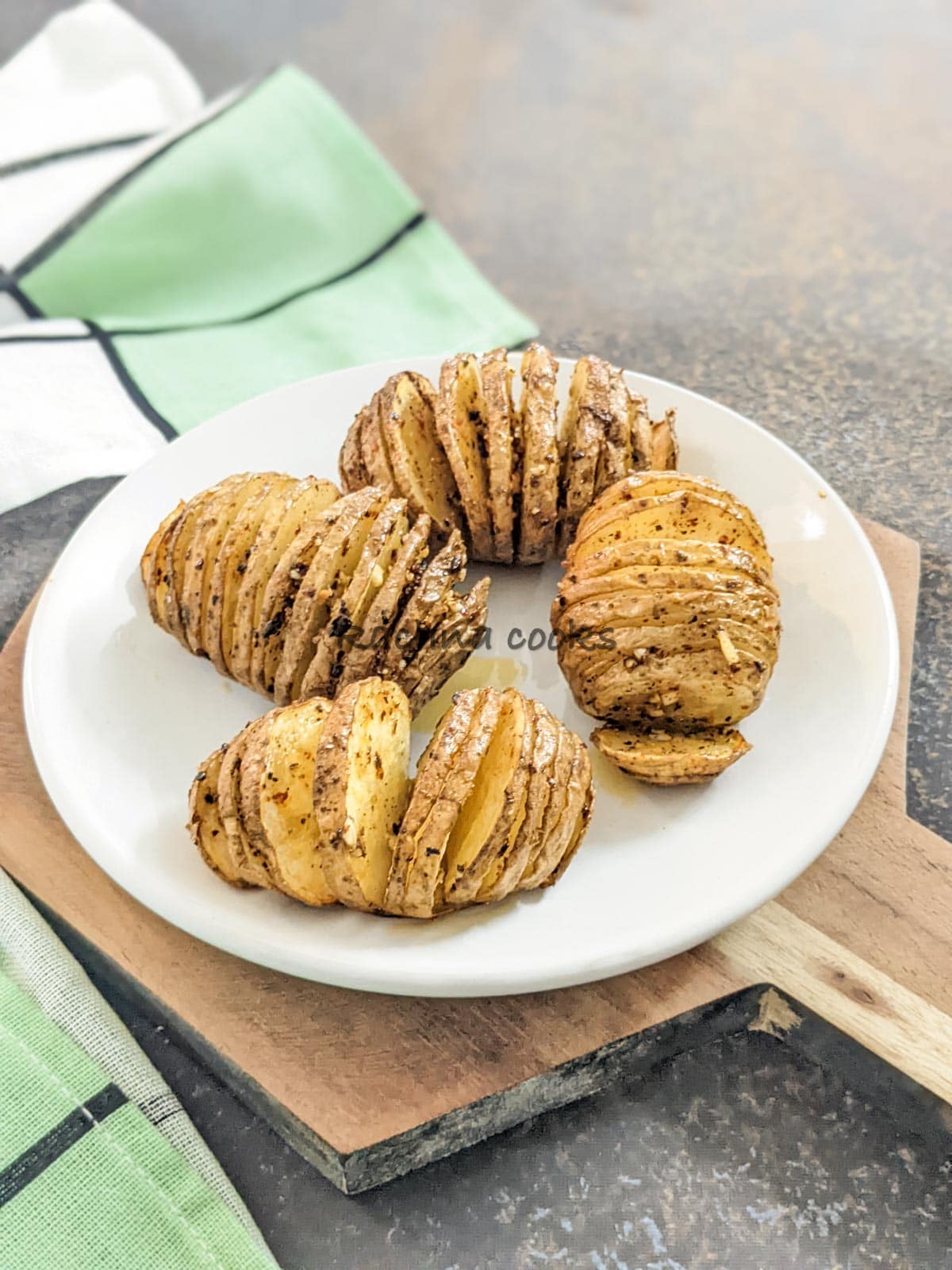 Four hasselback potatoes after air frying on a white plate with a green napkin on a brownish background.