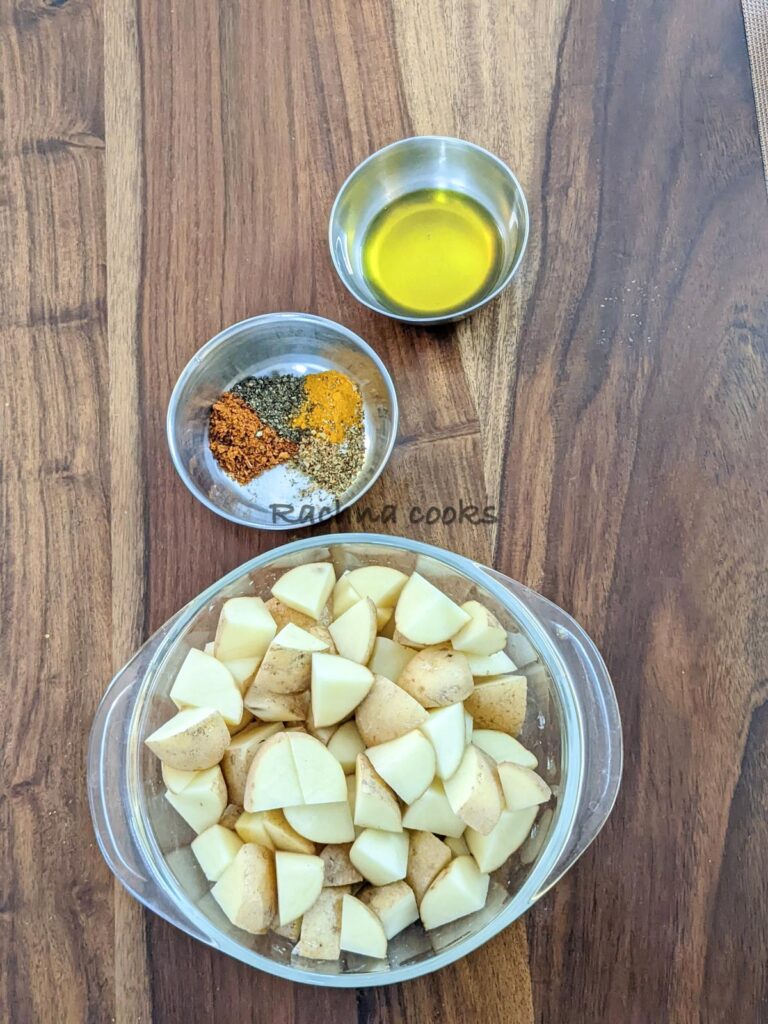 Peeled cut cubes of potatoes in a transparent bowl. One bowl of olive oil and one bowl with spices also in the picture.
