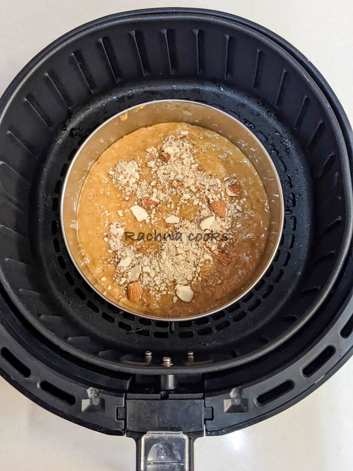 Cake tin with banana bread batter garnished with almond slivers is placed in air fryer basket ready for air frying.