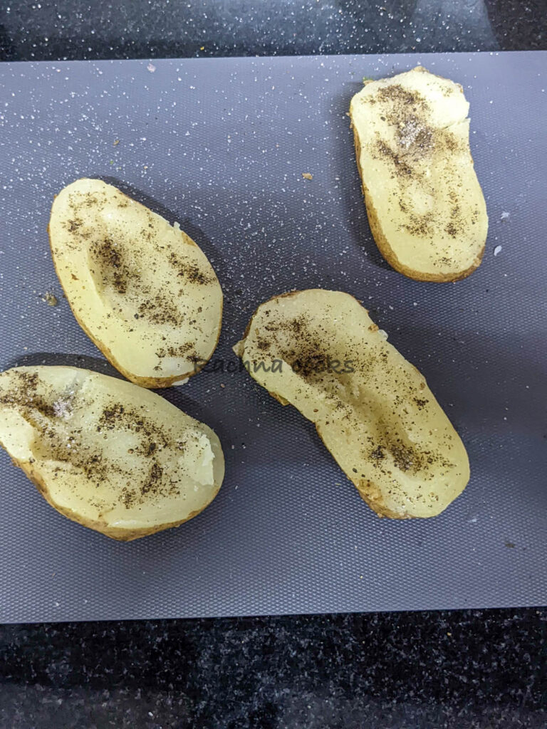 Potato boat halves on a grey chopping board with salt and pepper sprinkled on them.