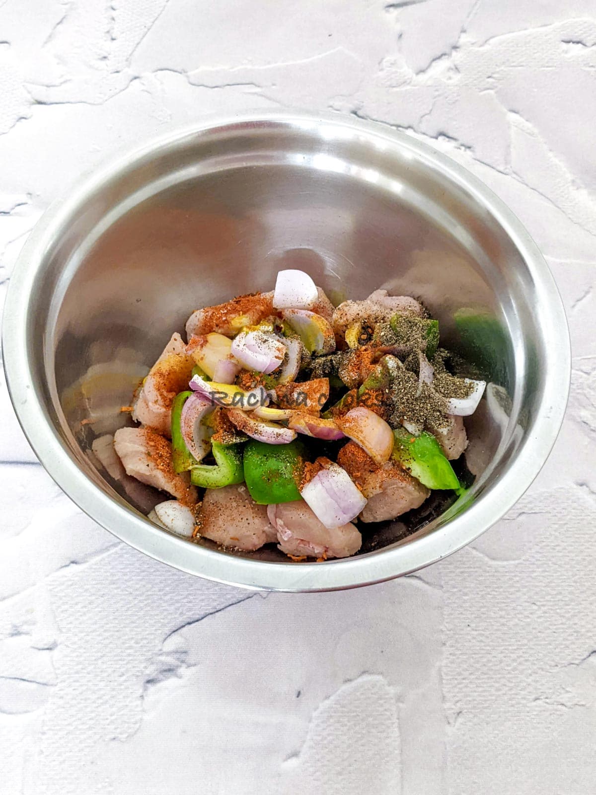 Spices and oil on top of peppers, onion and chicken cubes in a steel bowl.