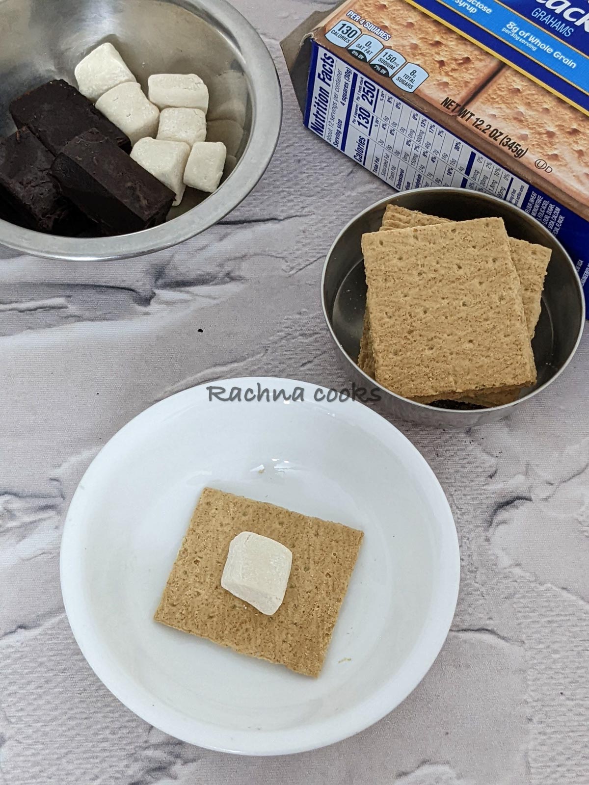 Graham's cracker with a pressed marshmallow on top.  Bowls of crackers, marshmallows and chocolate bars visible in the background.