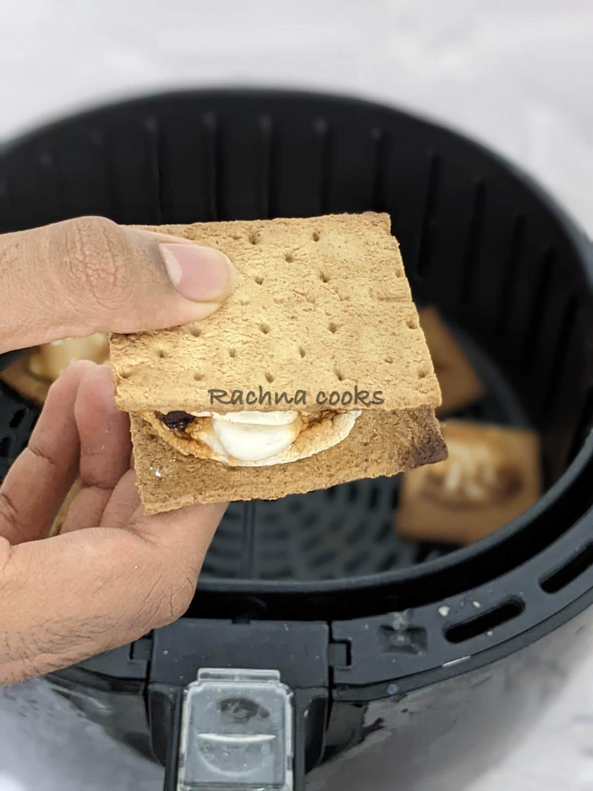 One s'more held in hand with the background of air fryer basket.