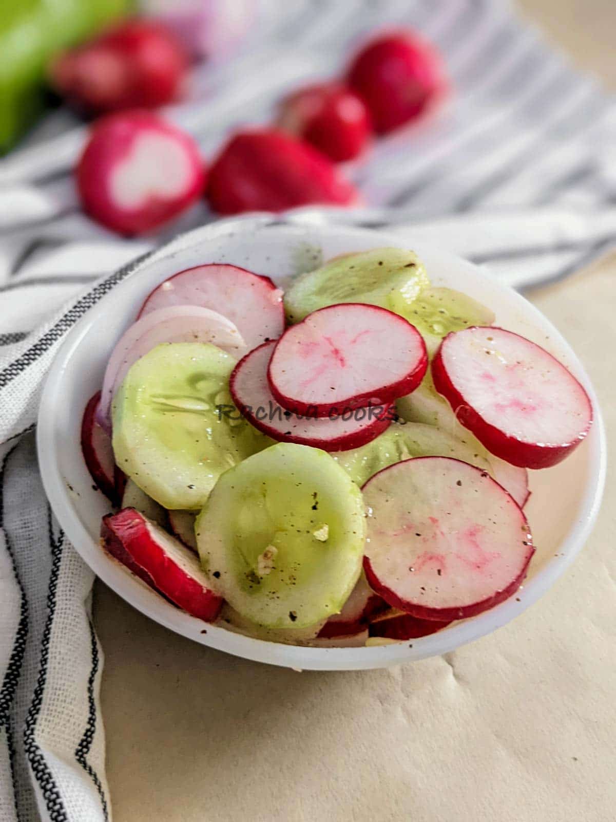 Delicious sliced radish, cucumber and onion salad with dressing in a white bowl with radishes, cucumber and onion in the background.