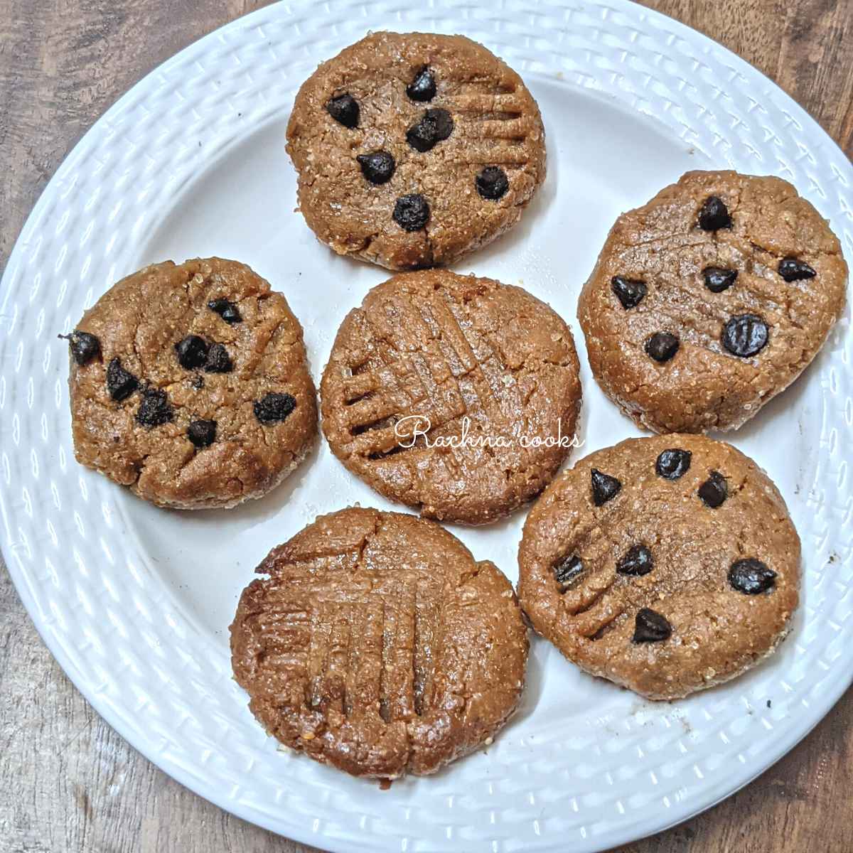 6 peanut butter cookies on a white plate, 2 of them having no chocolate chips and the rest with chocolate chips