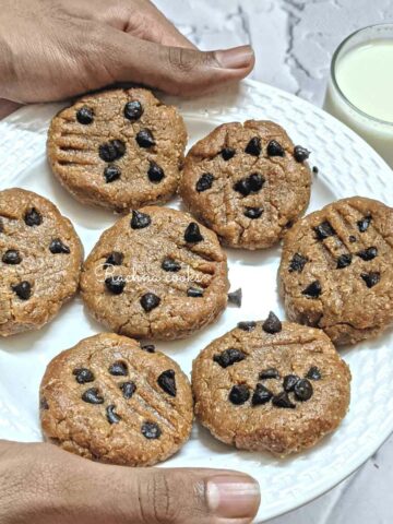 7 peanut butter chocolate chip cookies on a white plate held by hands with a glass of milk in the background.
