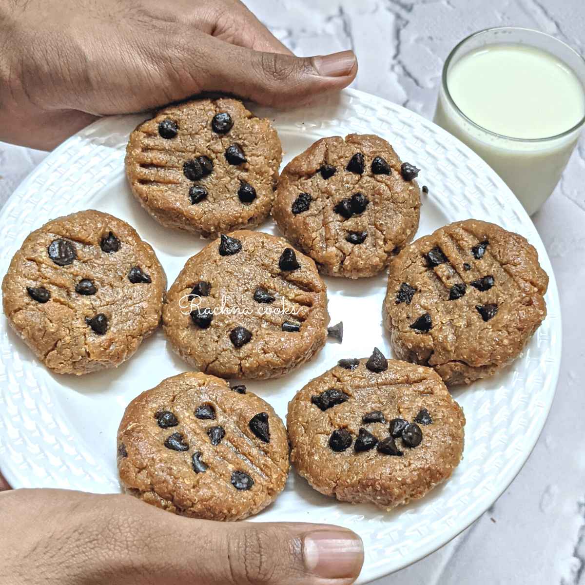7 peanut butter chocolate chip cookies on a white plate held by hands with a glass of milk in the background.