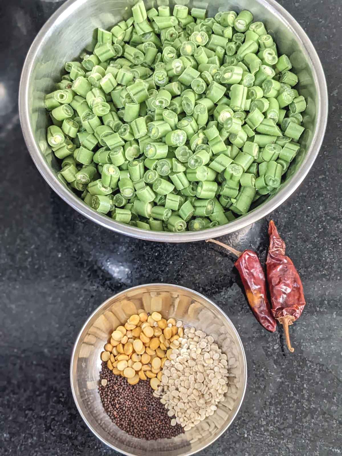 green beans cut into small pieces in a shallow bowl with lentils and mustard seeds in another bowl and 2 dry red chillis in the frame.