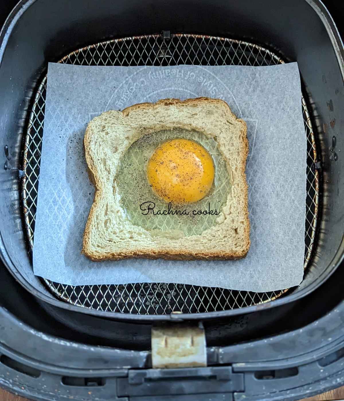 Bread with cavity cut out and with a broken egg with salt and pepper in air fryer basket.