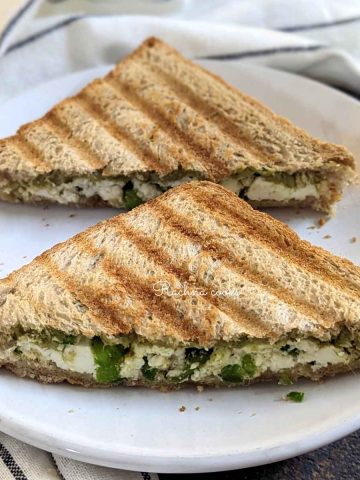 Two halves of one paneer sandwich cut into triangles served on a white plate.