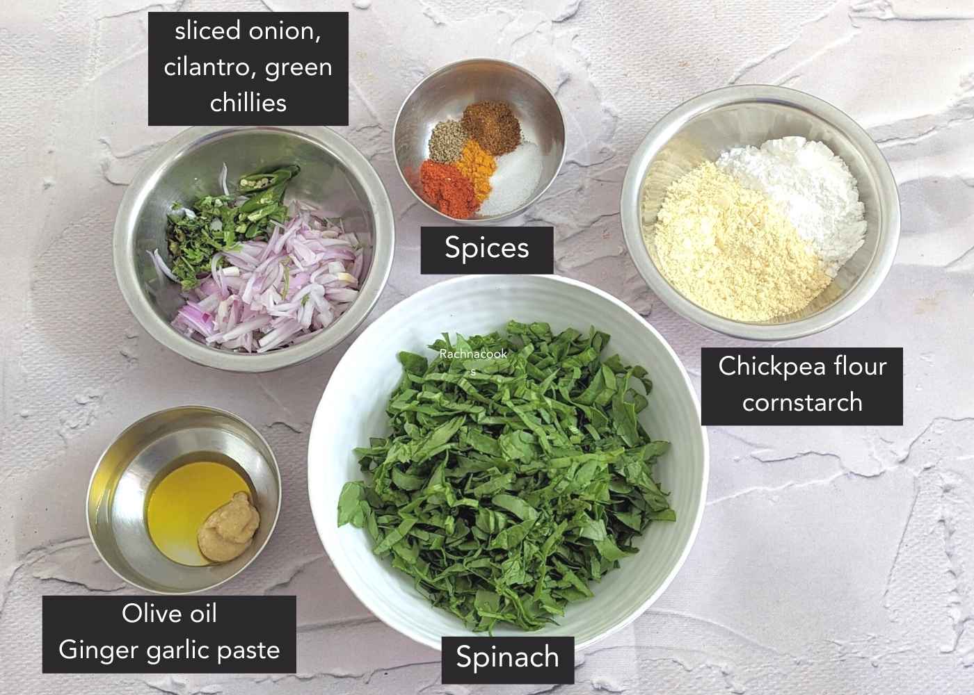 Ingredients for spinach pakora on a frame: spinach leaves, chopped onion, green chillies, cilantro, chickpea flour, cornstarch, spices, olive oil and ginger garlic paste in shallow bowls.