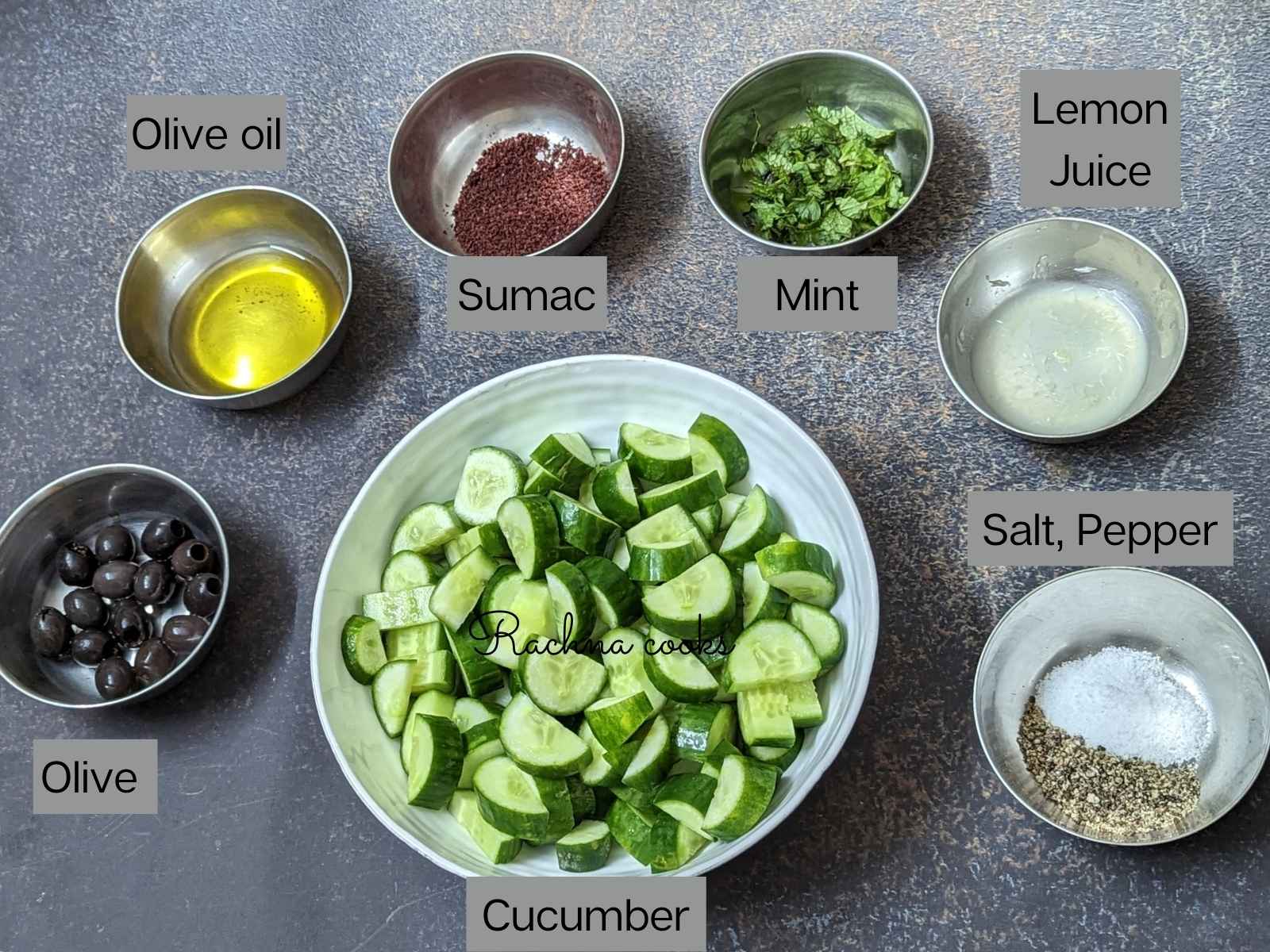 Ingredients for making sumac cucumber salad in bowls: Olive oil, chopped cucumbers, salt, pepper, sumac, mint, lemon juice and olives