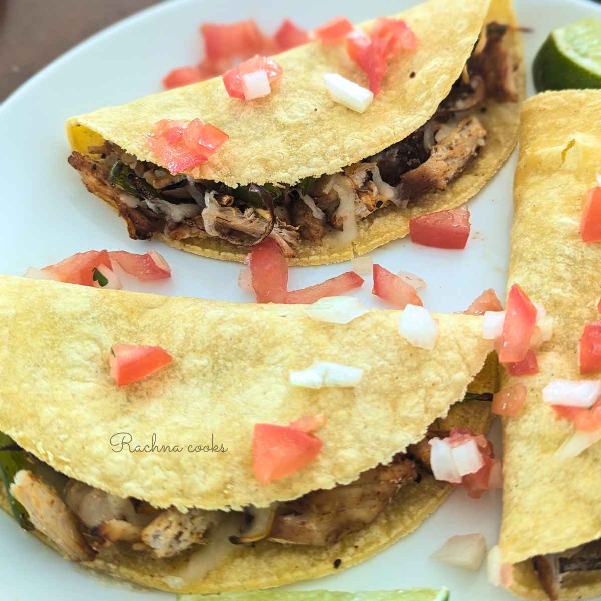3 chicken tacos served on a white plate with chopped onion, tomatoes and slices of lemon.